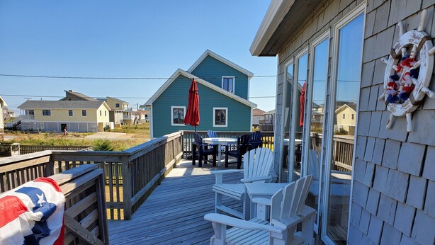 A wooden deck with white Adirondack chairs and a red umbrella, overlooking a beach with houses. - Steps to the Sand! Ocean Views! Hot tub! Specials! Dogs! KD0223 (Kill Devil Hills)