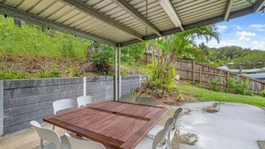 An outdoor patio with a wooden table and white chairs, surrounded by a wooden fence and lush greenery. - 17 Green Valley Drive - Rainbow Beach. Wifi. Walk to town and beach. (RAINBOW BEACH)