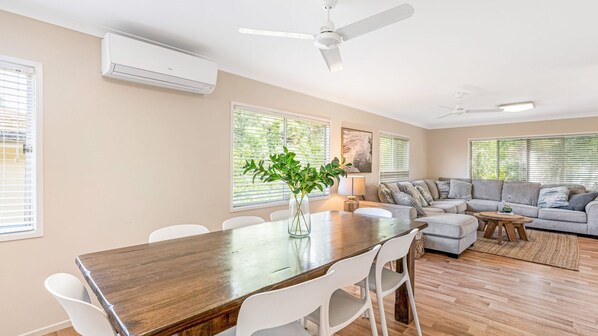 A spacious living room with a wooden dining table, white chairs, a grey sofa, and a ceiling fan. - 17 Green Valley Drive - Rainbow Beach. Wifi. Walk to town and beach. (RAINBOW BEACH)