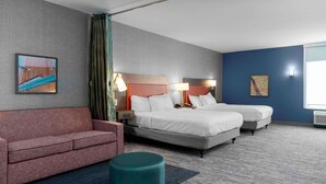 Quarto de hotel com duas camas, um sofá vermelho, um banquinho verde-azulado e obras de arte nas paredes.