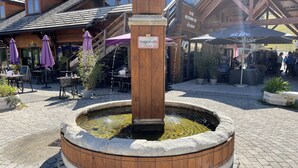Een houten fontein met een bordje, omringd door terrasstoelen en een restaurant op de achtergrond.