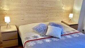 Een slaapkamer met een houten hoofdbord, een bed met een blauwe deken en witte sneeuwvlokkussens, en twee nachtlampjes op houten nachtkastjes.