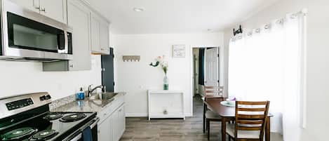 Una cocina moderna con armarios blancos, un microondas de acero inoxidable y una estufa. Hay una zona de comedor con una mesa y sillas de madera.