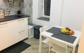 Een compacte keuken met witte kasten, een zwart aanrecht, een raam en een eethoek met een witte tafel en stoelen.