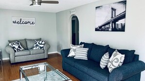 Ett vardagsrum med en grå soffa, en blå soffa och ett soffbord i glas. Det finns en takfläkt, en välkomstskylt och en inramad tavla på väggen.