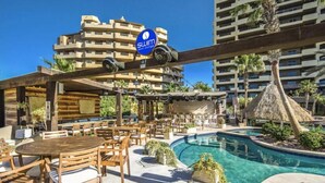 Un espace aménagé au bord de la piscine, avec des tables et des chaises en bois, un panneau indiquant l’accès à un espace de détente, et une piscine entourée de palmiers et d’une structure à toit de chaume.