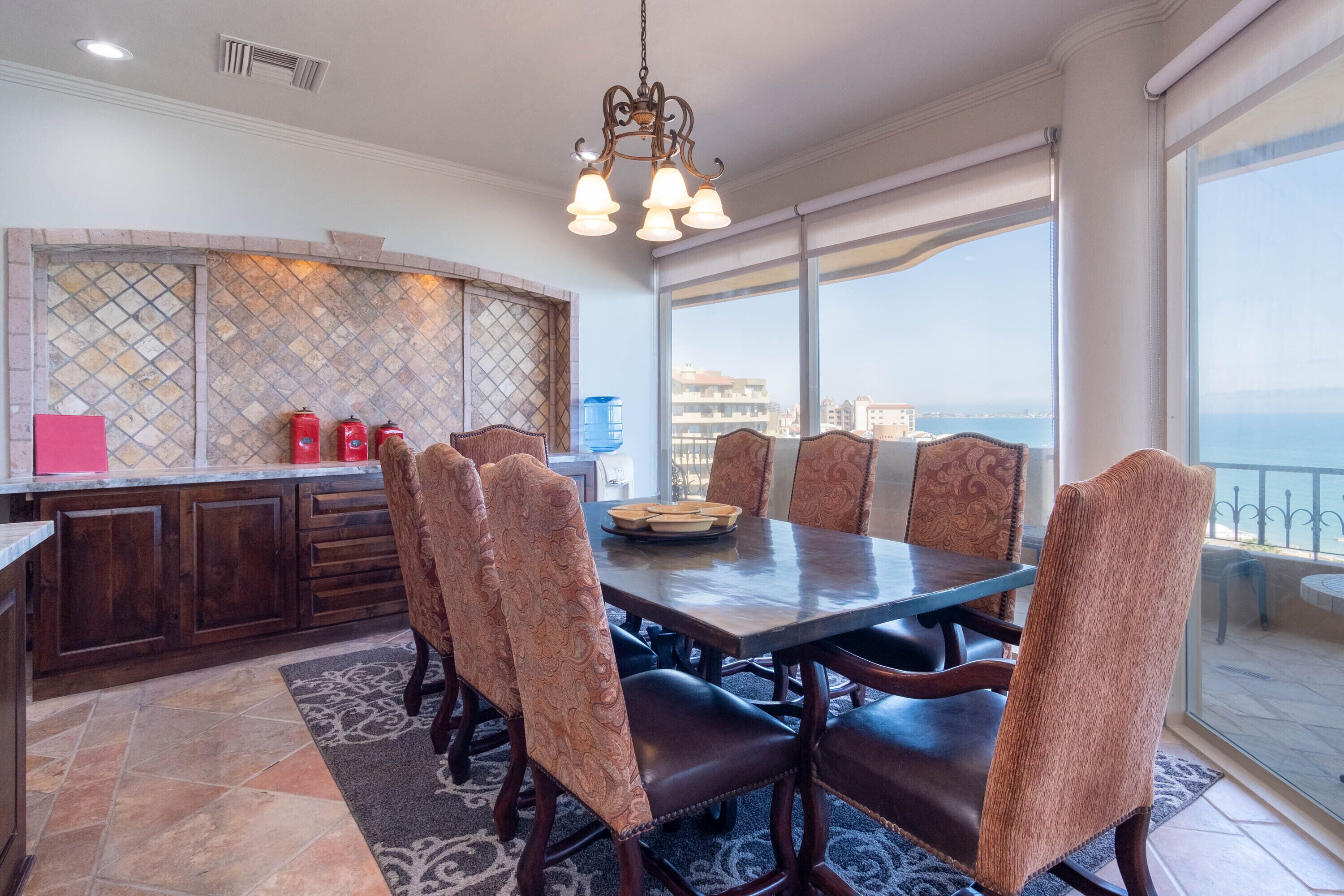Un espace repas avec une grande table et des chaises à motifs, un mur décoratif et une vue sur la mer.