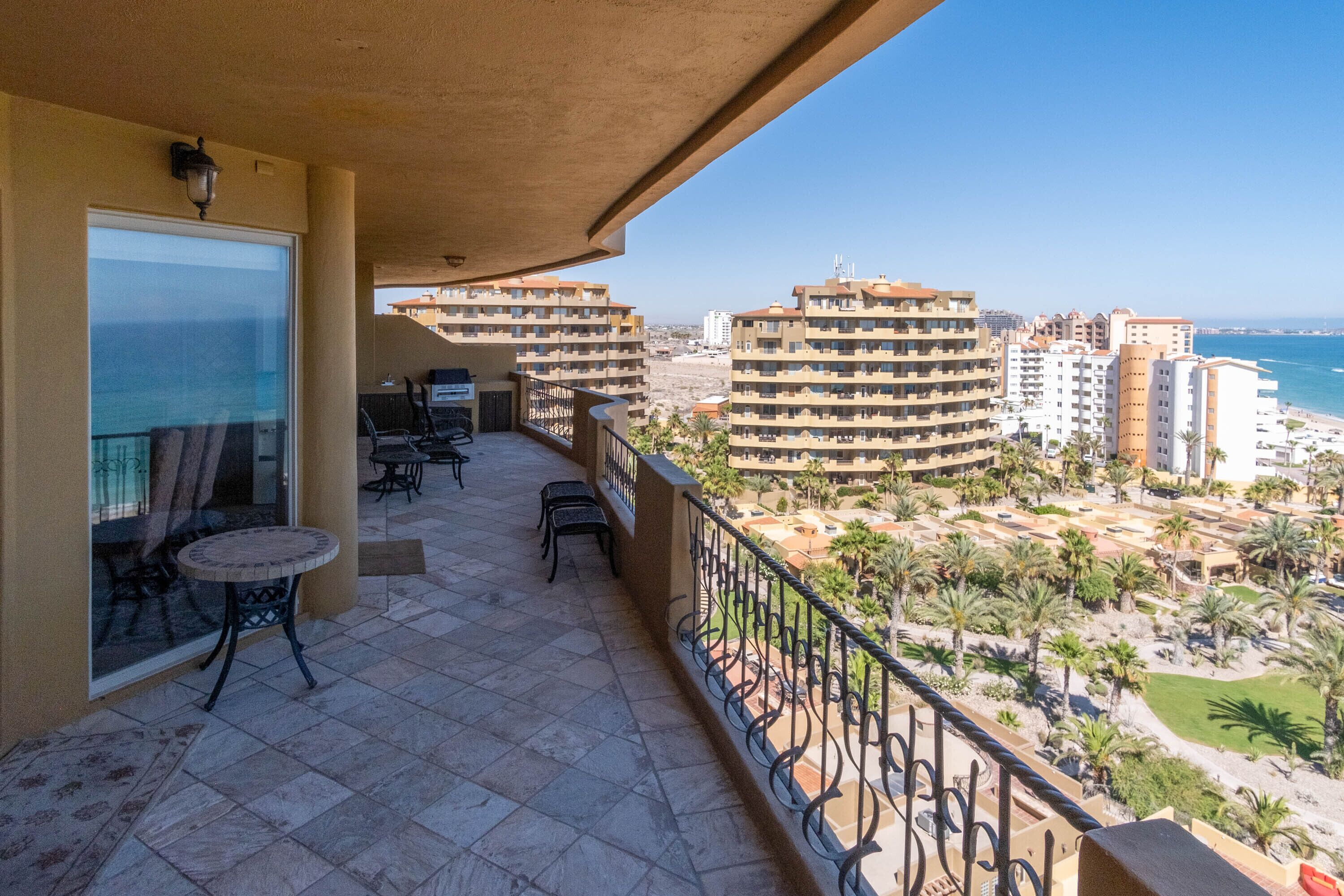 A balcony with outdoor seating, a view of the sea, and surrounding buildings.