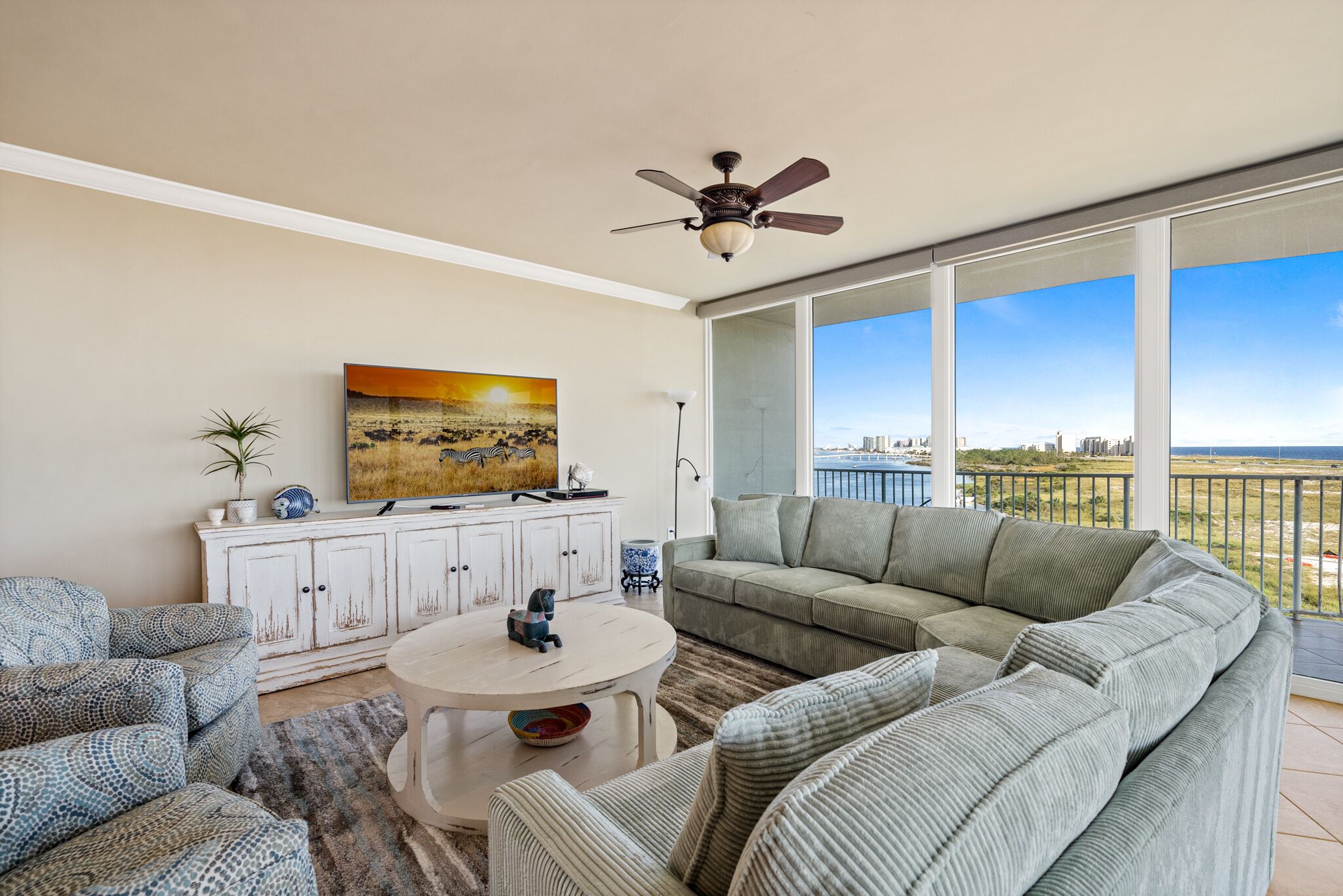A spacious living room with a large gray sectional sofa, a round coffee table, and a ceiling fan. A painting and a cabinet are on the wall.