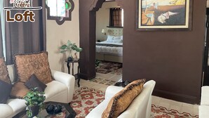 A cozy living room with a sofa, armchairs, a coffee table, and a small side table with a plant. A framed painting hangs on the wall, and a bed is visible in the background. - La Casa del Colibrí (Campeche)