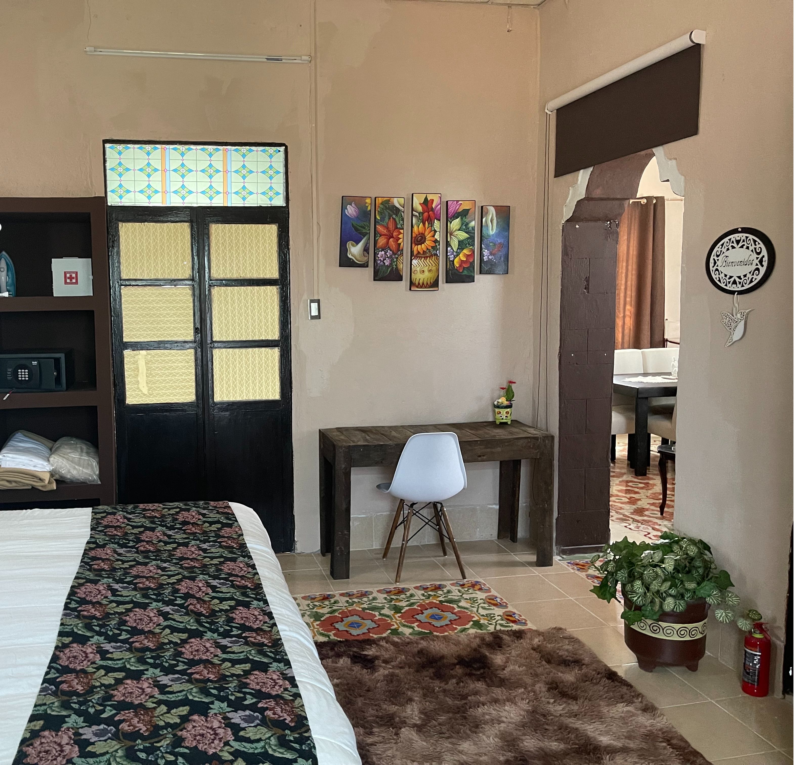 Une chambre d’hôtel avec un lit, un bureau, une chaise et une porte agrémentée d’un paravent décoratif. On y trouve également des œuvres d’art murales et un panneau d’accueil.
