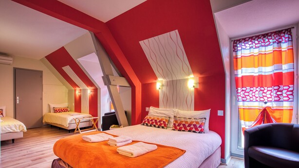 A hotel room with a sloped ceiling, a large bed with an orange blanket, a chair, and a window with striped curtains.