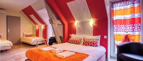 A hotel room with a sloped ceiling, a large bed with an orange blanket, a chair, and a window with striped curtains.