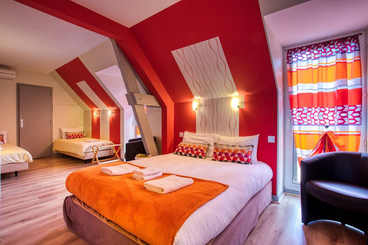 A hotel room with a sloped ceiling, a large bed with an orange blanket, a chair, and a window with striped curtains.