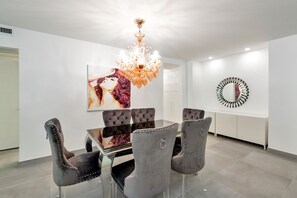 Uma sala de jantar moderna com mesa de vidro, cadeiras estofadas cinzentas, um candelabro e duas obras de arte na parede.