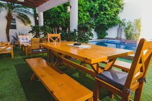 Un área de comedor al aire libre con una mesa y bancos de madera, rodeada de abundante follaje y una piscina.