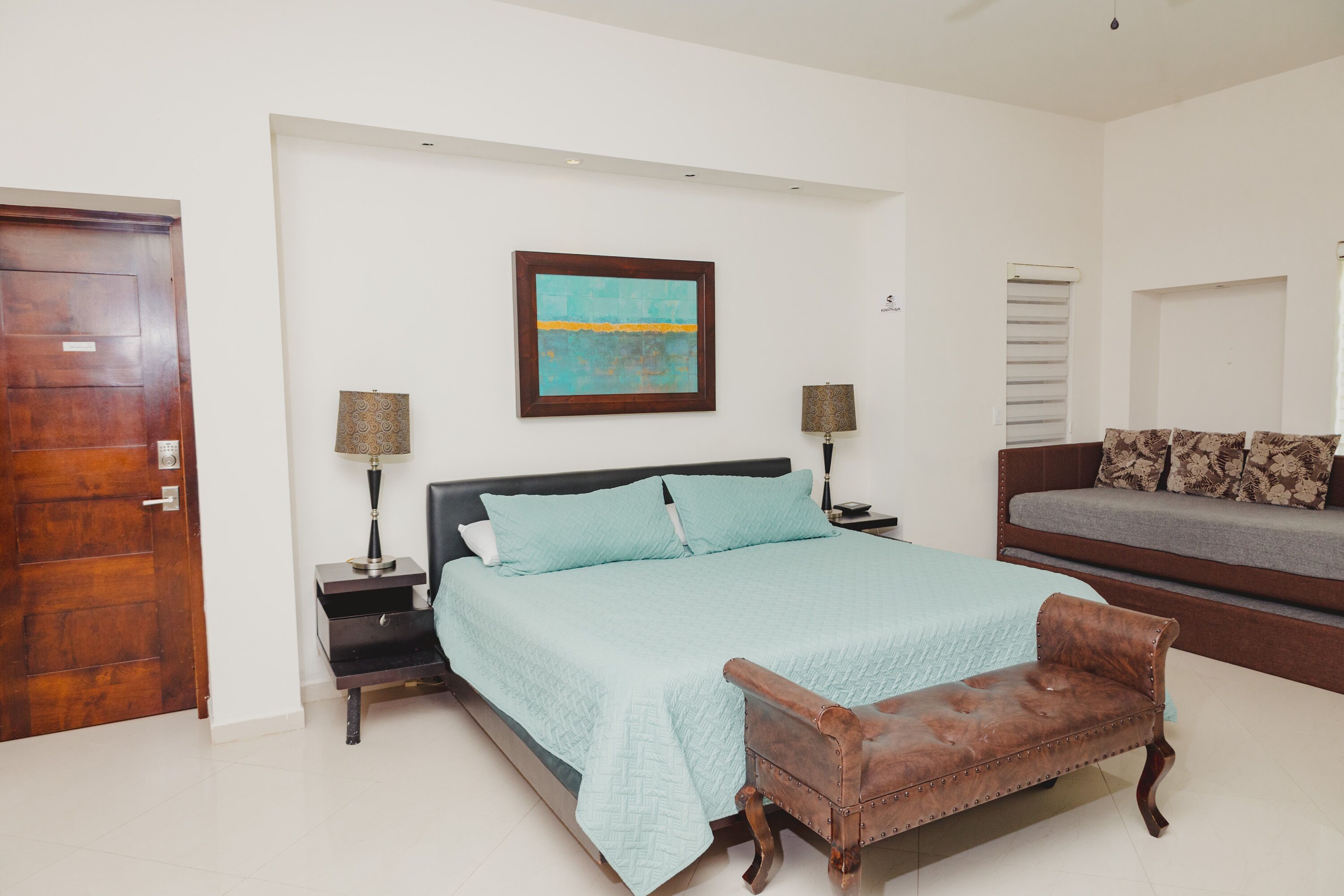 A bedroom with a bed, bedside tables, a bench, a sofa, and a painting on the wall.