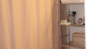 A bathroom with a beige shower curtain, a metal shelf with toiletries, and a toilet. - Cozy Chic Loft (Detroit)