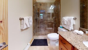 Un baño con ducha de vidrio, inodoro blanco, encimera de granito y alfombra negra.
