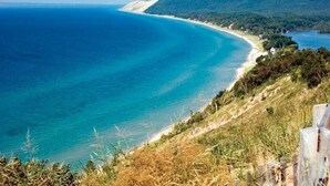 A coastal landscape with a beach, forested hills, and a clear view of the ocean. - Glen Den Amazing Views of Sleeping Bear Dunes, Luxury Lakefront Home Glen Arbor (Empire)
