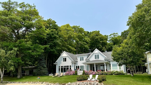 A two-story house with a large front porch, surrounded by lush greenery and a sandy beach. - Glen Den Amazing Views of Sleeping Bear Dunes, Luxury Lakefront Home Glen Arbor (Empire)