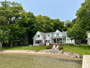 Une maison à deux étages dotée d’un grand porche à l’avant, entourée d’une végétation luxuriante et bordée d’une plage de sable.