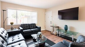 A modern living room with a black leather sofa, a glass coffee table, and a flat-screen TV mounted on the wall.