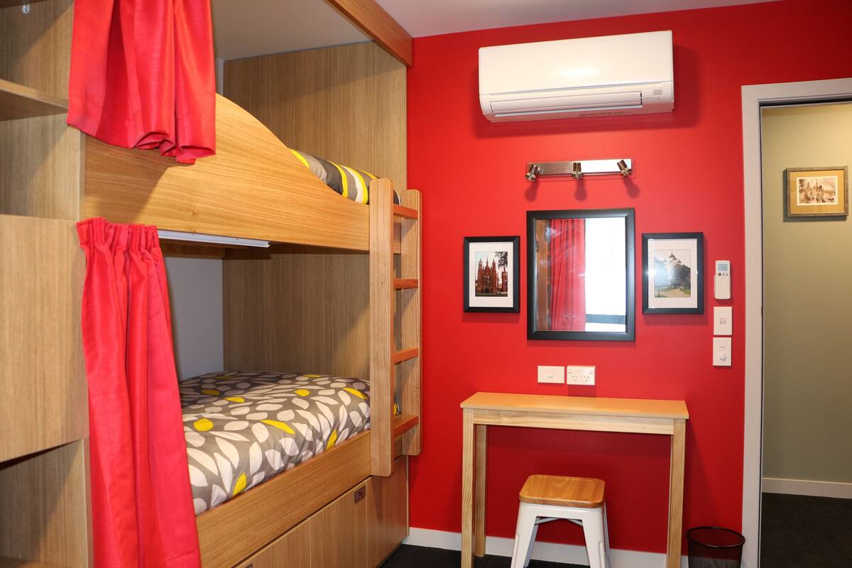 Una litera con cortina roja, una mesita pequeña y un aire acondicionado instalado en la pared.