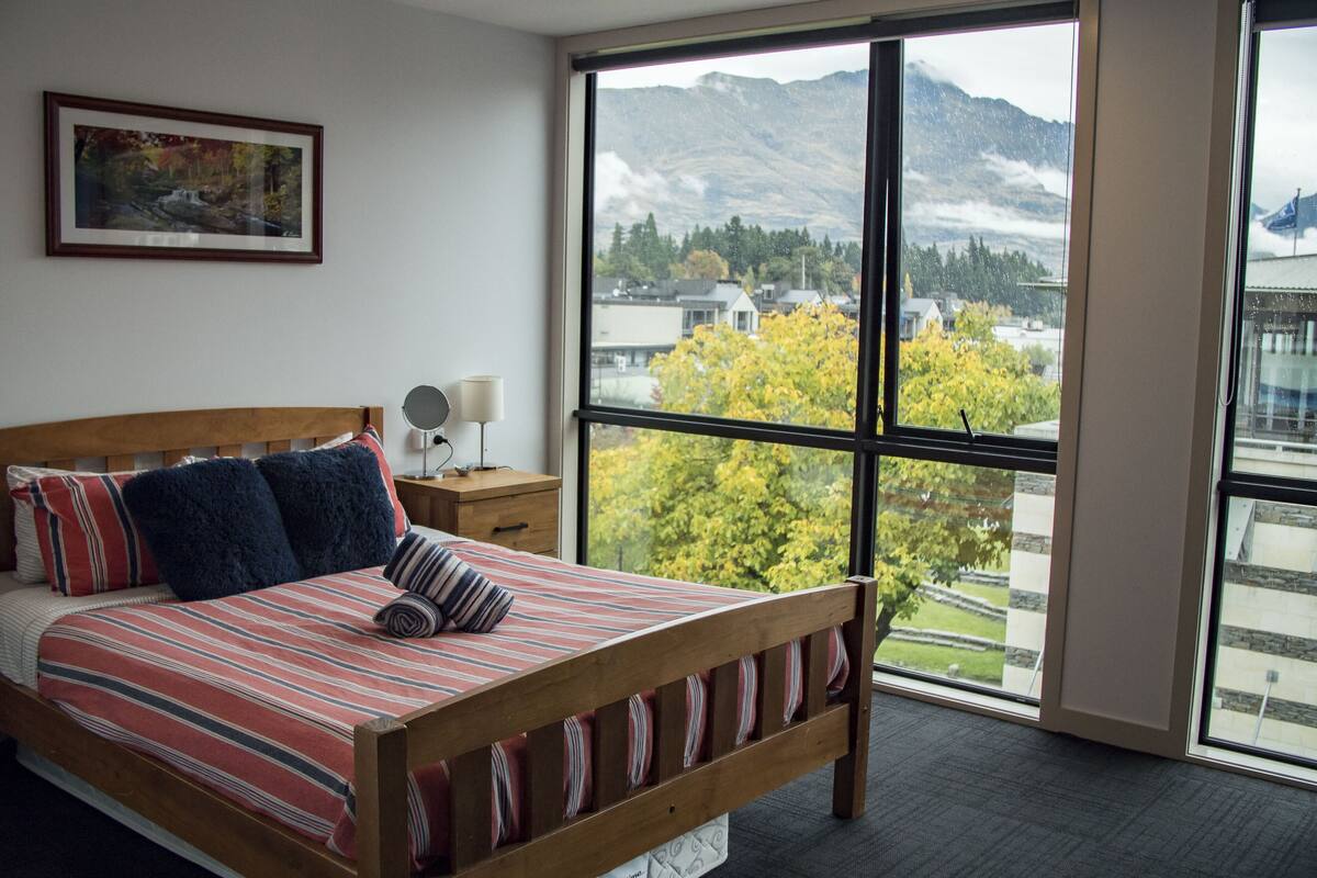 Un dormitorio con un ventanal grande, una cama de madera, una mesita de noche con una lámpara y un cuadro en la pared.
