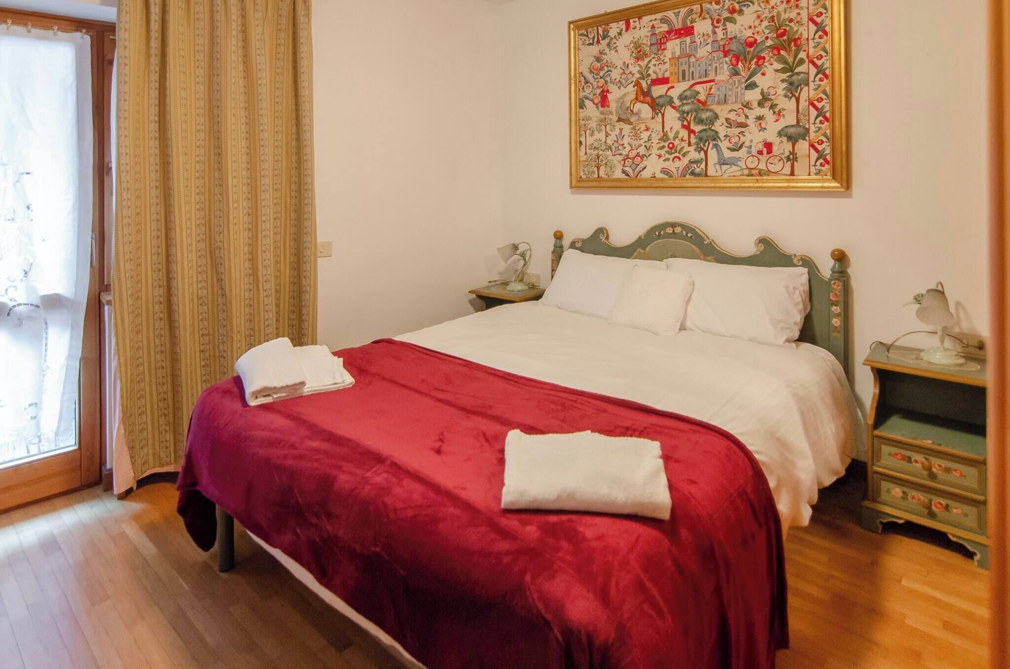 A neatly made bed with a red blanket and white pillows, a wooden nightstand with a lamp, and a floral painting on the wall.