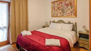 Un letto rifatto con una coperta rossa e cuscini bianchi, un comodino in legno con una lampada e un quadro floreale sulla parete.