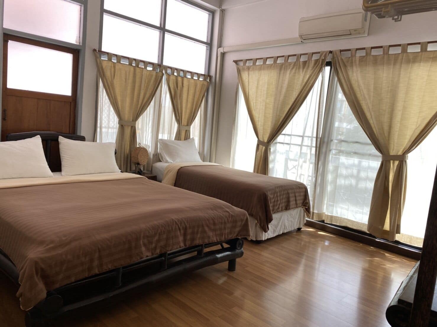 A hotel room with two beds, beige curtains, wooden flooring, and an air conditioning unit.