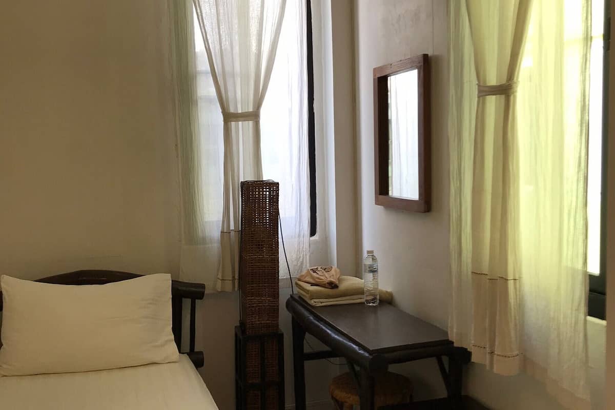 Ein Hotelzimmer mit Bett, Schreibtisch, Stuhl, Hocker, Spiegel und leichtem Vorhang.