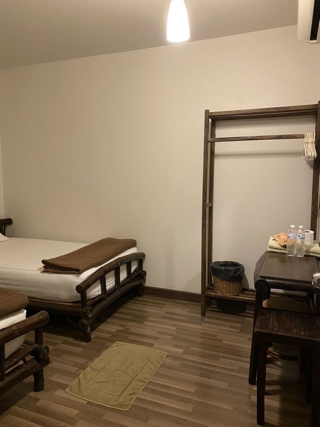 A room with a bed, a wooden desk, a chair, a small table, and a trash bin.