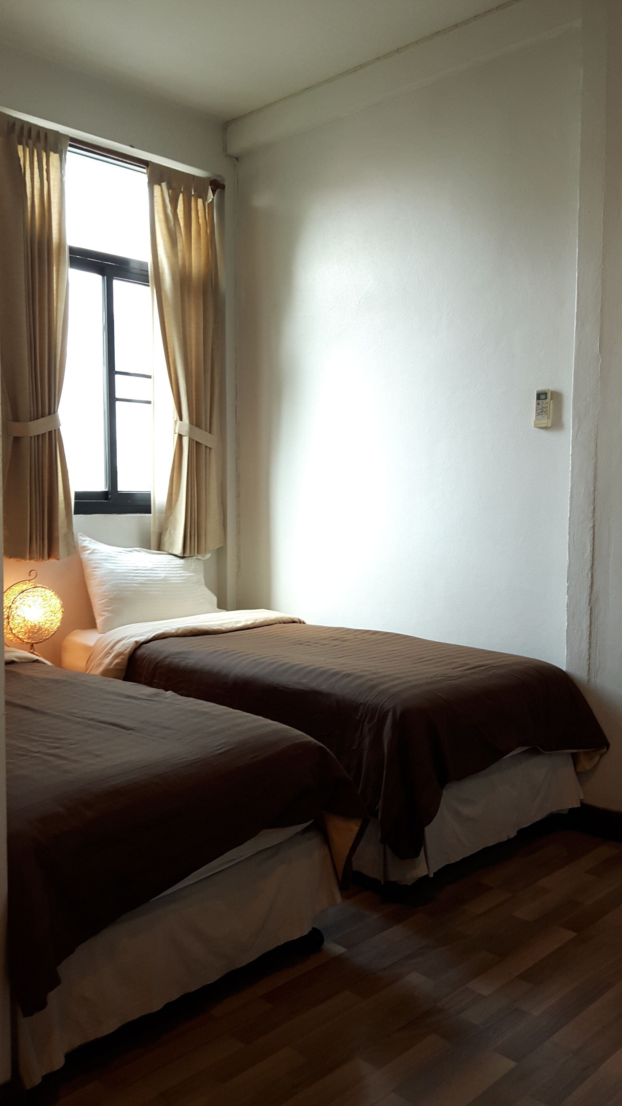 A bedroom with two beds, a window with curtains, a bedside lamp, and a wall-mounted phone.