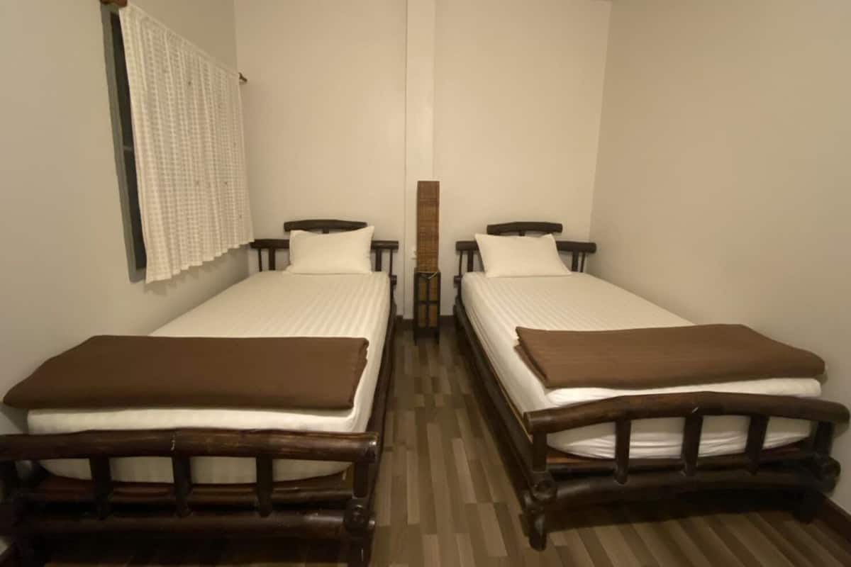 Ein Zimmer mit zwei Einzelbetten, jeweils mit Holzrahmen und weißer Bettwäsche. An der linken Seite befindet sich ein Fenster mit einem weißen Vorhang.