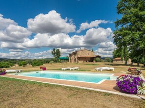 Uma área de piscina com um edifício de pedra, mobiliário para áreas externas e vegetação ao redor.