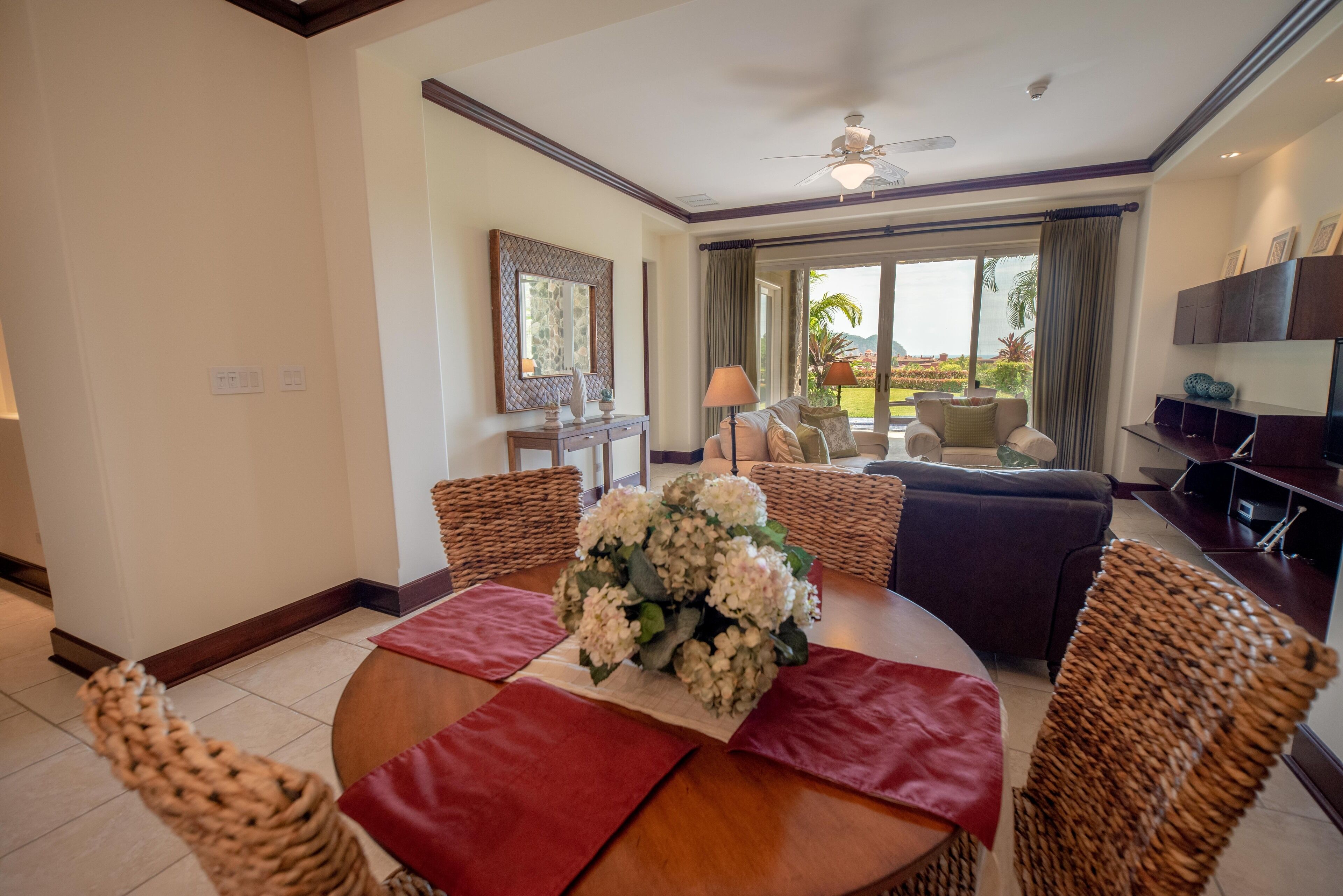 Uma sala de jantar com uma mesa redonda, cadeiras de vime e um centro de mesa floral. Há um sofá e uma televisão ao fundo.