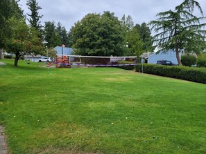Zona exterior amb gespa, una xarxa de voleibol, una petita zona de jocs i un edifici blau al fons.