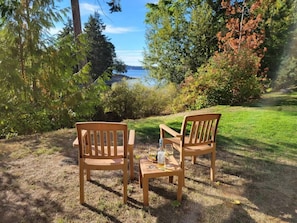 Two wooden chairs and a small table with a bottle and glasses in a grassy outdoor area with trees and a view of a body of water.