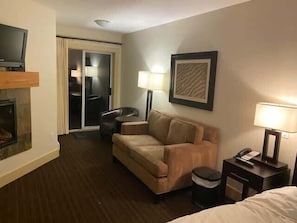 A hotel room with a sofa, a small table with a lamp, a television, and a fireplace.