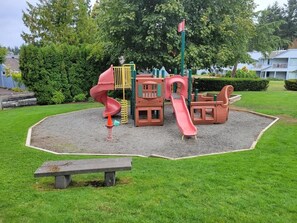 A playground with slides and climbing structures, surrounded by a grassy area and trees.