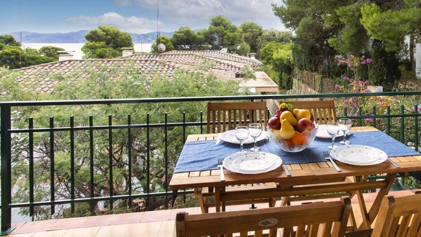 Une table en bois, dressée pour deux personnes, avec des assiettes, des couverts et un panier de fruits, sur un balcon donnant sur un paysage pittoresque, avec des arbres et une vue lointaine sur des montagnes.
