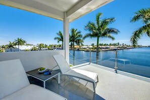 A balcony with white lounge chairs, a small table with a bowl of fruit, and a view of the marina and palm trees.
