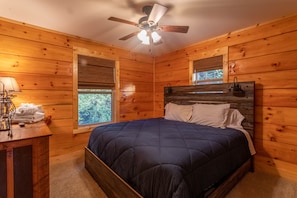 Une chambre à coucher dans une cabane en bois, avec un lit, un ventilateur de plafond, une table de chevet et une fenêtre avec des stores.