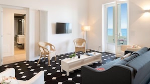 Un salon moderne avec un sol carrelé en noir et blanc, un téléviseur à écran plat fixé au mur, un canapé, deux fauteuils en osier, une table basse blanche et une vue sur la mer par une grande fenêtre.