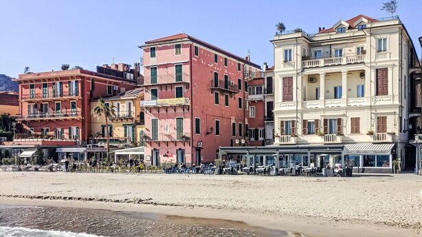Un hôtel en bord de mer, avec plusieurs étages, des espaces de détente en plein air et une plage de sable au premier plan.