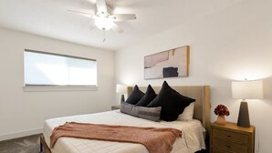 Un dormitorio con cama, mesita de noche, lámpara, ventilador de techo, ventana y un cuadro en la pared.