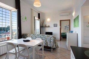 A room with a dining table, chairs, a bed with a floral bedspread, a small TV, and a refrigerator.