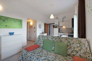 Un espace de vie comprenant un canapé, une commode blanche, une œuvre d’art murale verte et une cuisine en arrière-plan.